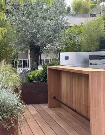 Une jardinière courbe en acier Corten, pleine de végétation, posée sur un parquet. Des graminées, un olivier. La cuisine d'extérieur au premier plan.