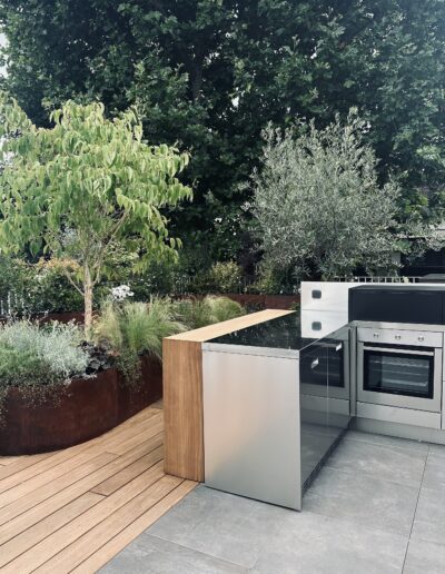 Une jardinière courbe en acier Corten, pleine de végétation, posée sur un parquet. Des graminées, un olivier. La cuisine d'extérieur au premier plan.