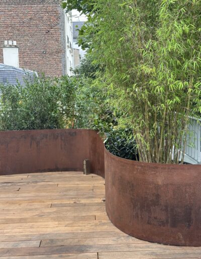 Une jardinière courbe en acier Corten, pleine de végétation, posée sur un parquet. Des bambous.